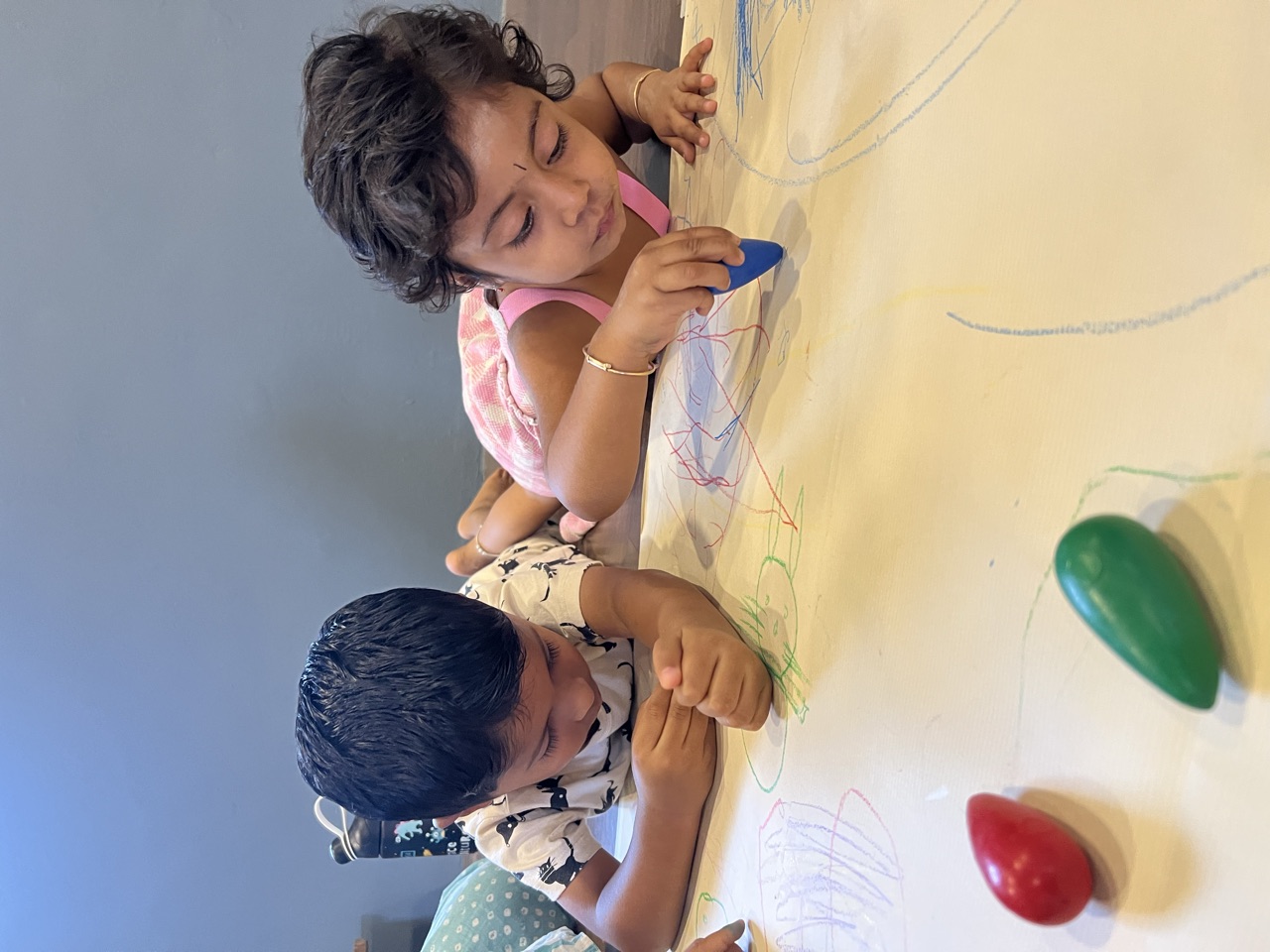 Two children drawing together with crayons on a big shared sheet of paper