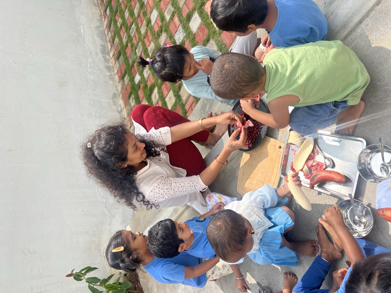 Children around a teacher opening a pomegranate over trays of fresh fruit