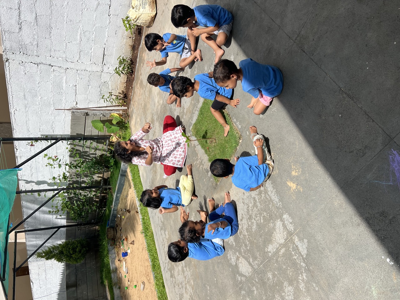 Children sitting in a circle around a young plant with their teacher in the courtyard