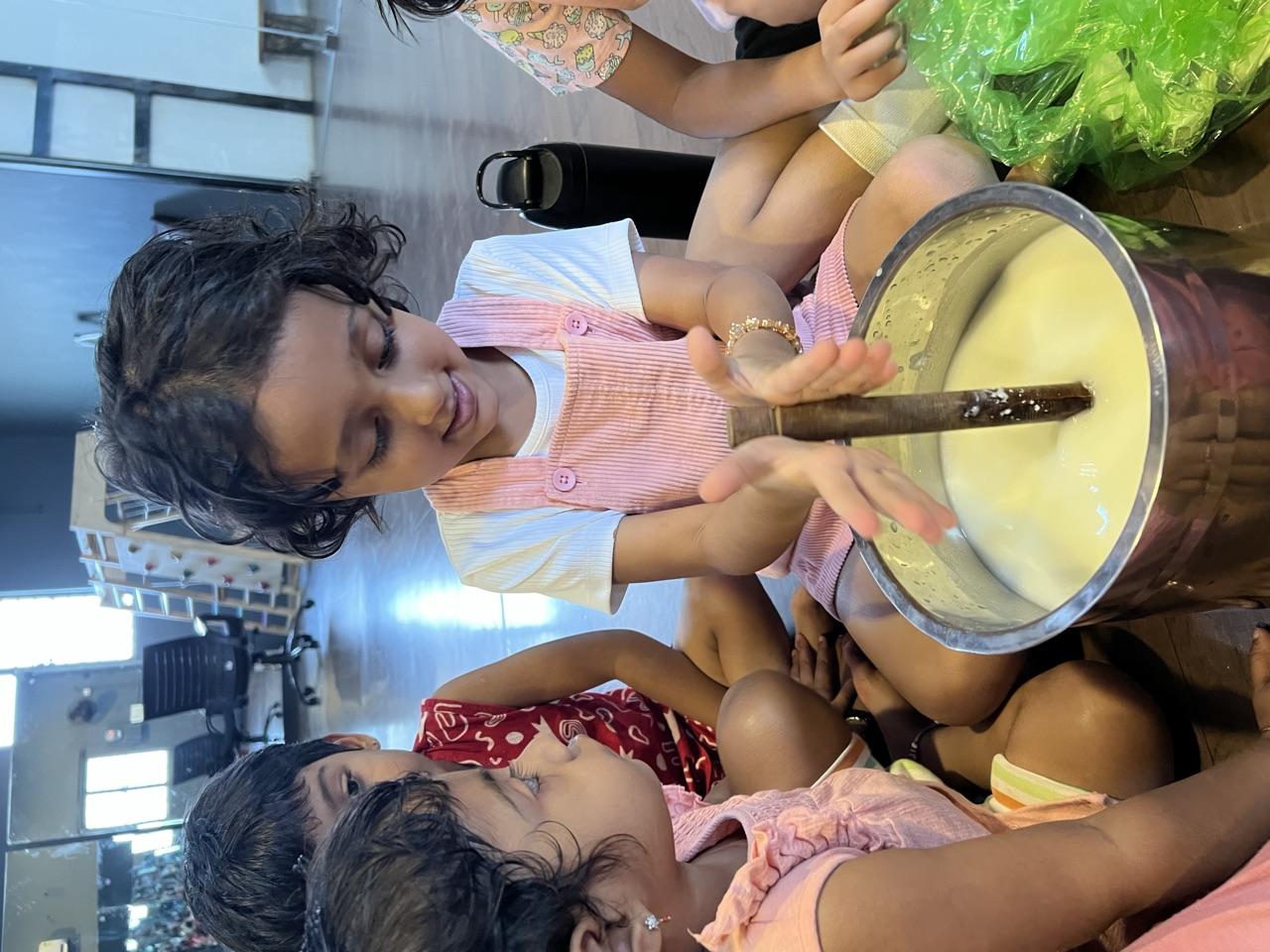 A child churning a big pot of milk with a wooden churner while friends watch